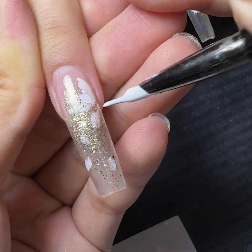 Un pennello liner fine che dipinge piccoli petali di fiori bianchi su ununghia trasparente con glitter oro.
