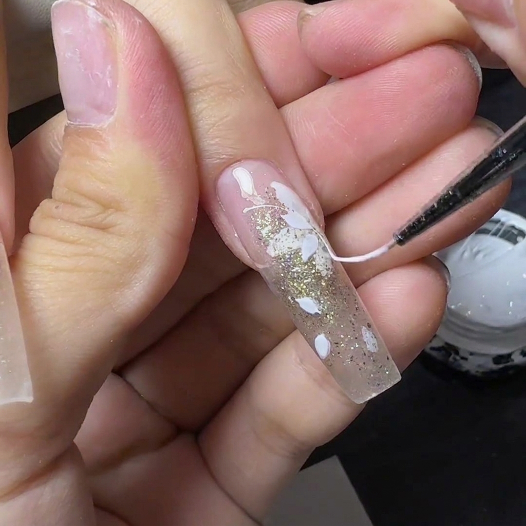 Uno strumento sottile che tira gel ragnatela bianco filamentoso attraverso ununghia decorata con fiori bianchi e glitter oro.
