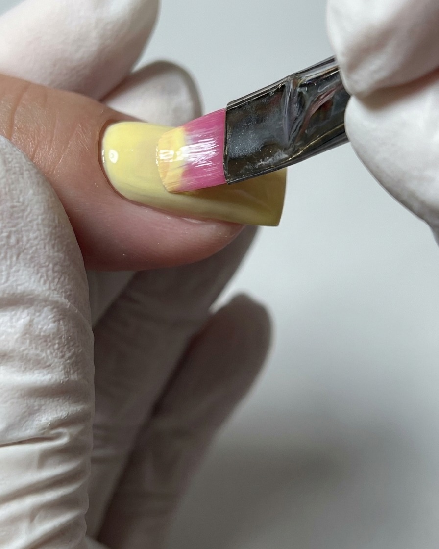 Un primo piano di un pennello che applica un secondo strato di smalto gel giallo pastello per garantire una copertura completa del colore.