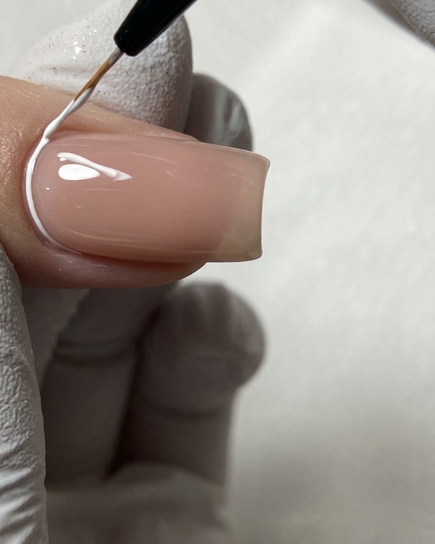 Un pennello fine per nail art che disegna linee geometriche bianche vicino alla cuticola su ununghia nude.