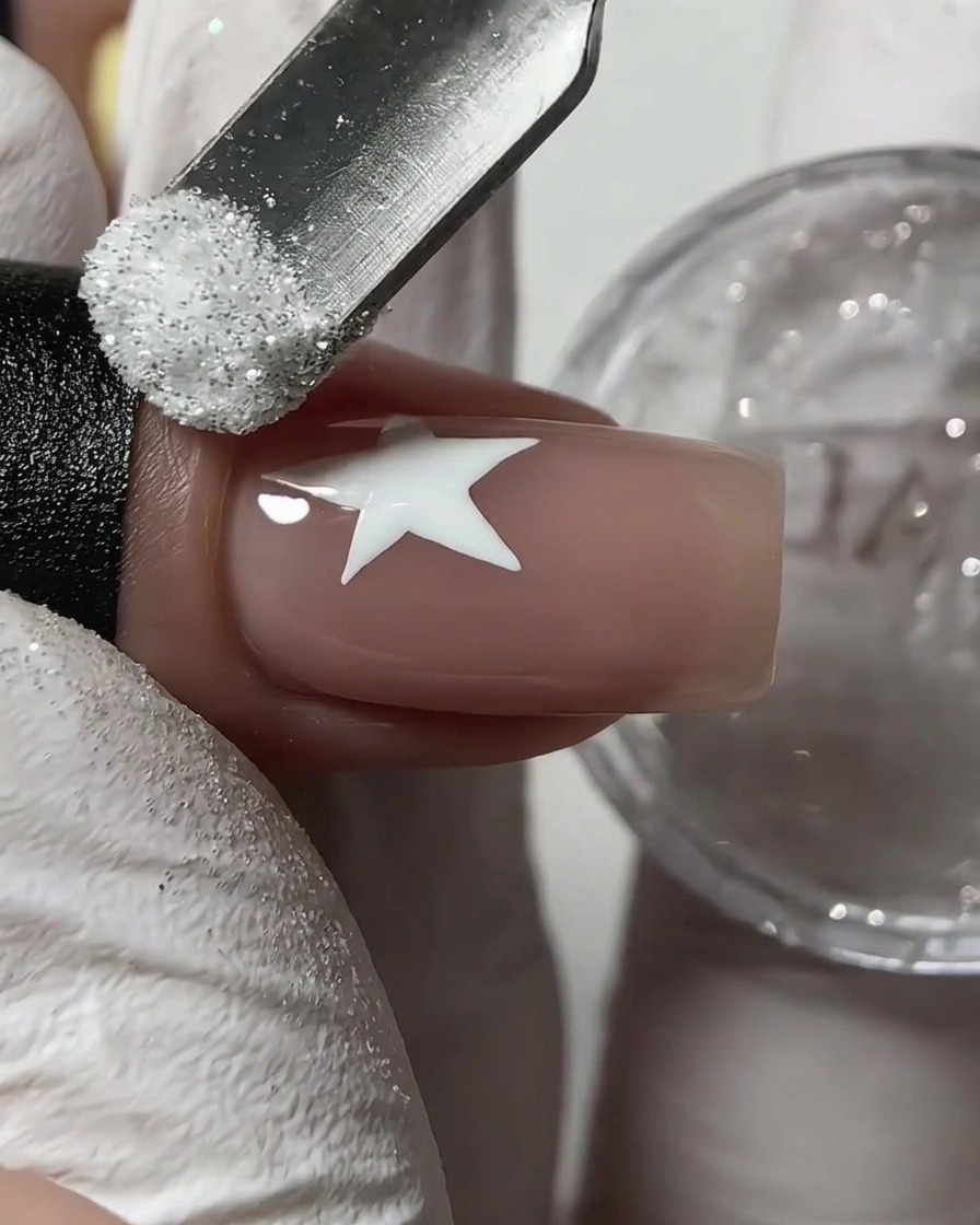 Uno spingicuticole in metallo che cosparge polvere glitter zucchero bianca su una stella bianca bagnata disegnata su ununghia nude.