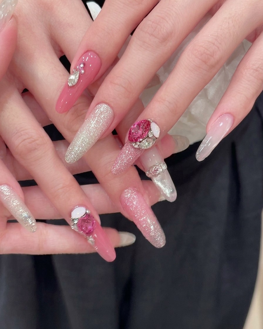 Due mani perfettamente curate che riposano su uno sfondo di tessuto scuro, mostrando unghie lunghe rosa a mandorla rifinite con gradienti di glitter argento e grandi accenti di strass a cuore rosa.