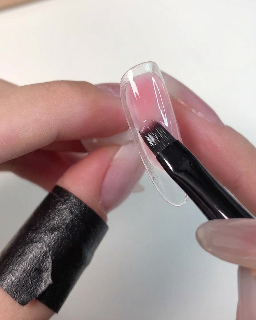 Una mano che tiene una tip trasparente mentre un pennello piatto tampona e sfuma delicatamente il gel rosa bagnato per ammorbidirne i bordi.