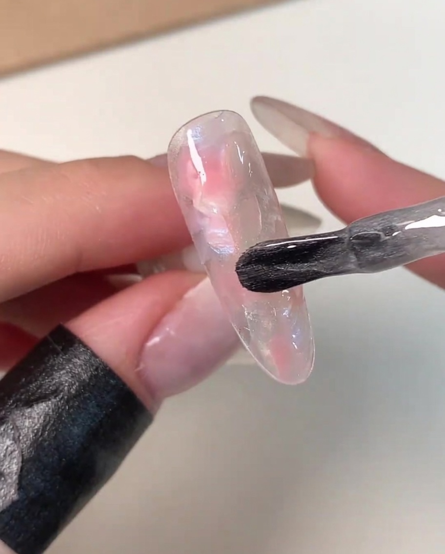 Un pennello da boccetta che applica una mano di gel magnetico trasparente e brillante sopra una tip decorata con effetto marmo rosa e bianco.