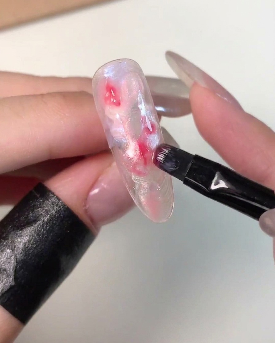 Un piccolo pennello piatto che applica gocce di gel rosa più scuro al centro di una tip con effetto marmo non polimerizzata.