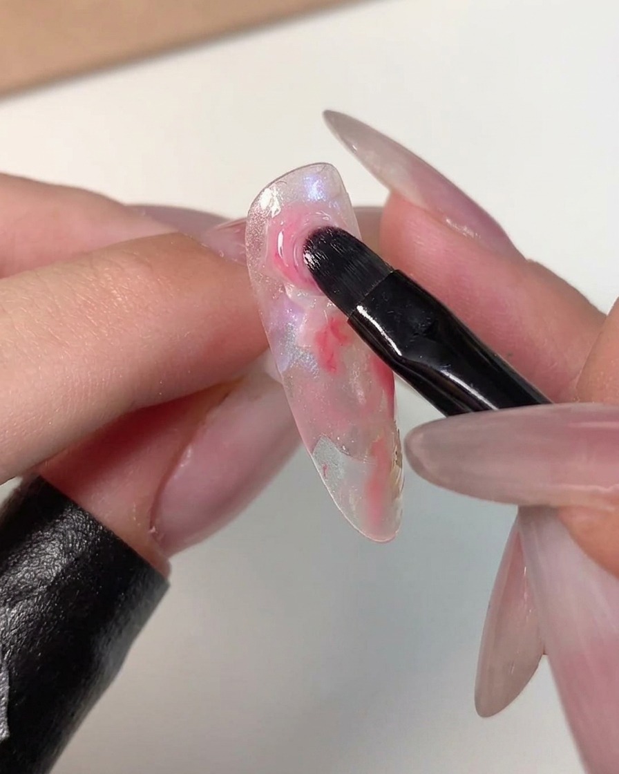 Un pennello da nail art che sfuma i bordi di un punto rosa scuro nei colori più chiari circostanti su una tip per unghie trasparente.