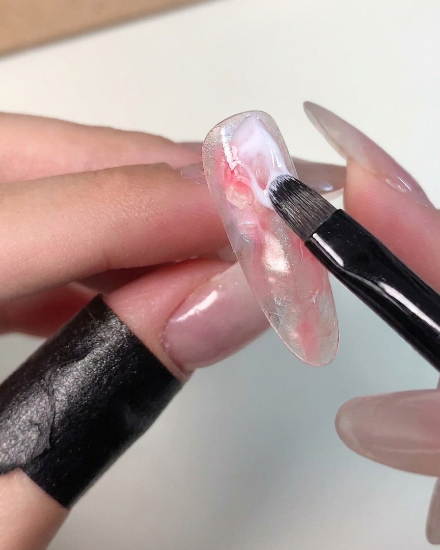 Un piccolo pennello da gel che applica una pennellata di gel bianco latte vicino allarea delle cuticole di una tip marmorizzata per perfezionare il motivo.