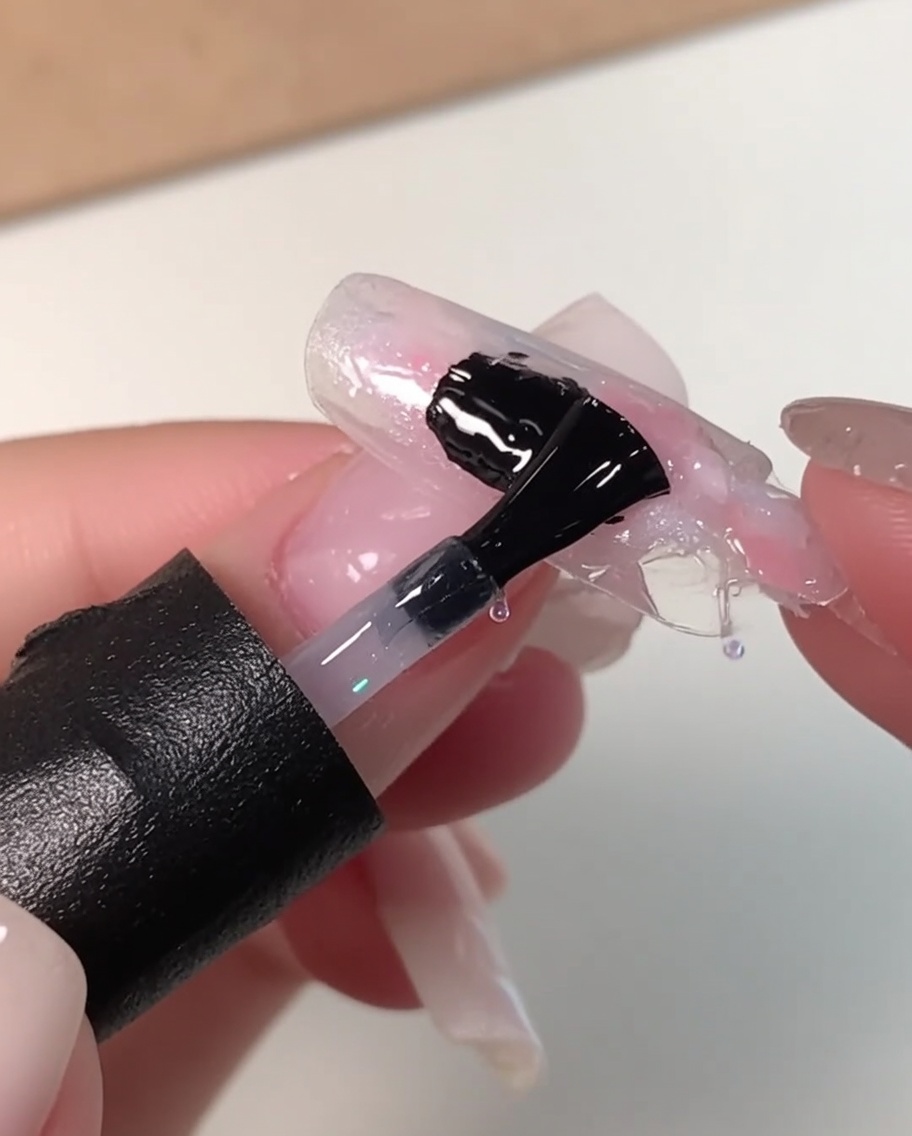 Un pennello per unghie nero che applica uno strato spesso di gel livellante trasparente su un design per unghie con sfumature rosa, bianche e blu.