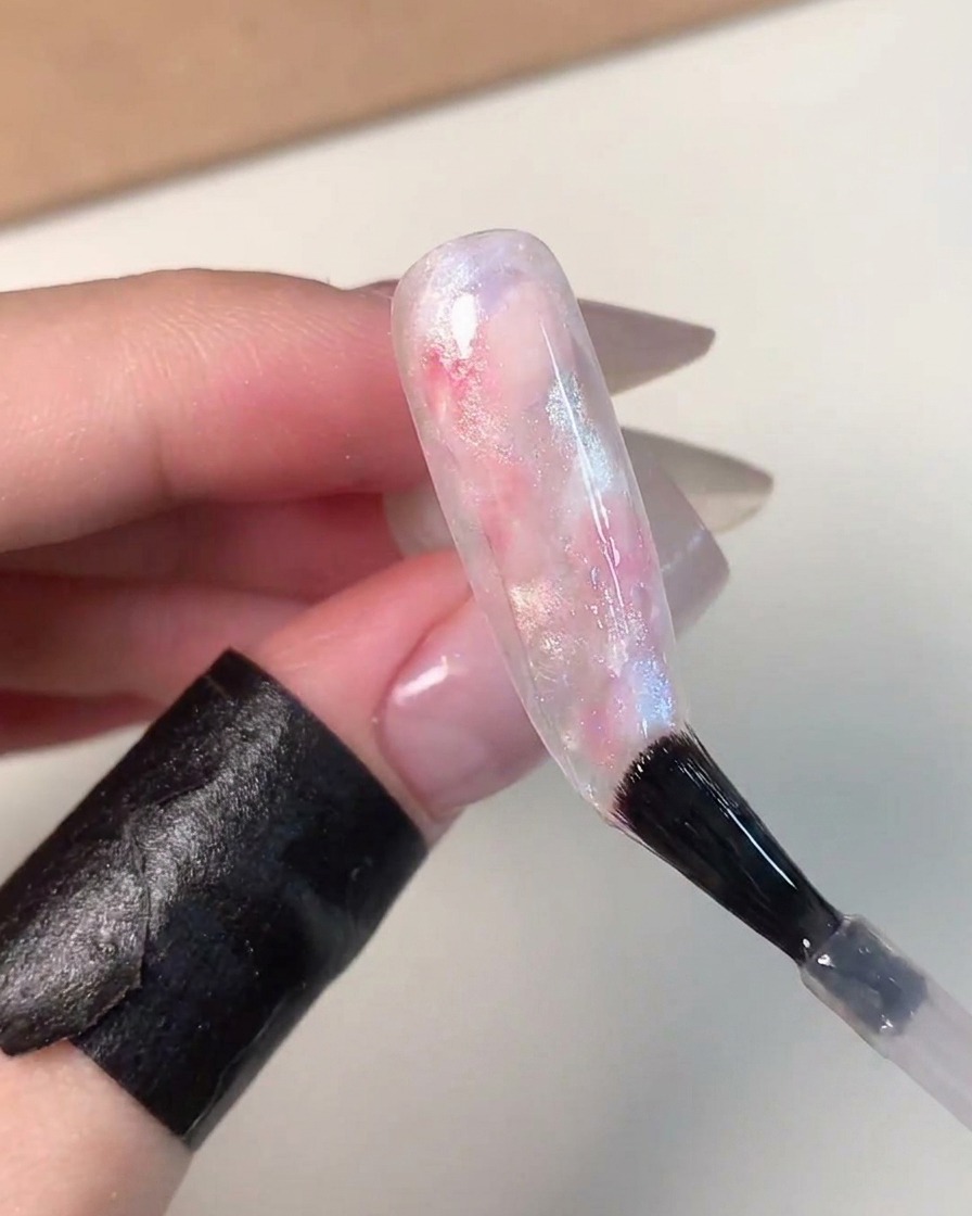 Un pennello da unghie che applica un top coat trasparente e lucido su ununghia finita con effetto sfera di vetro e sfumature rosa, blu e bianche.