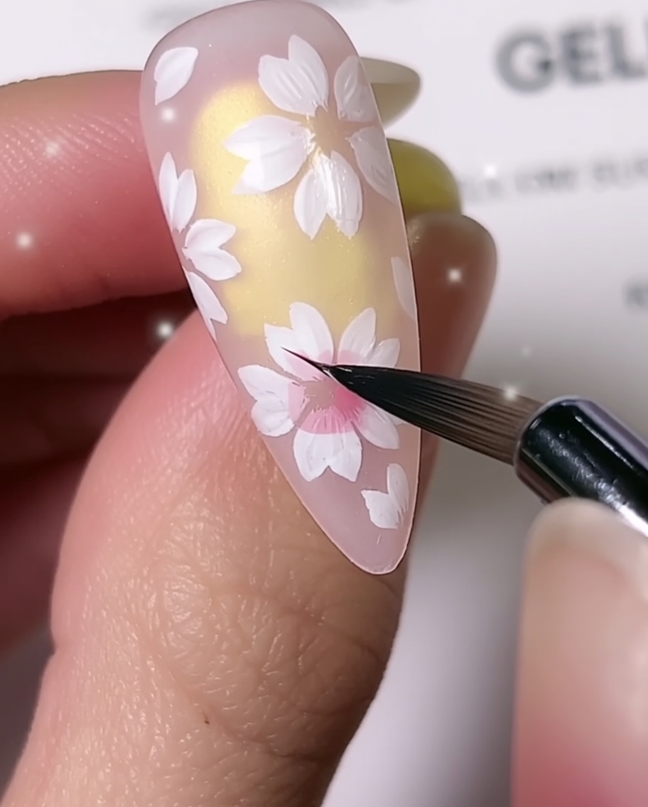 Un primo piano che mostra un pennello per dettagli fini che aggiunge ombreggiatura rosa al centro dei petali di ciliegio bianchi su ununghia a mandorla.