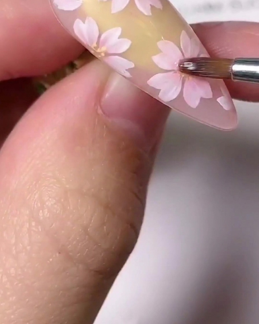 Un primo piano di un pennello per dettagli fini che applica piccoli accenti dorati al centro di un fiore di ciliegio rosa dipinto su ununghia.