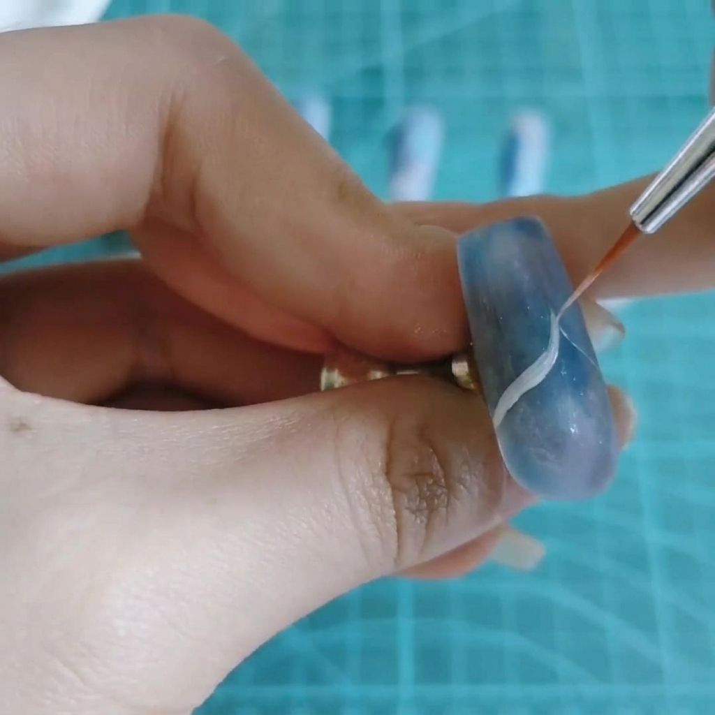 Un pennello fine per nail art traccia una sottile linea ondulata bianca su ununghia marmorizzata blu e viola.