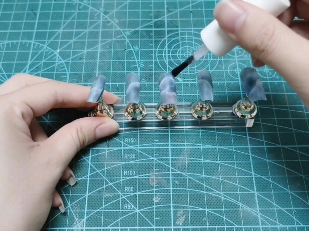 Un pennello a boccetta applica del gel costruttore trasparente su un set di unghie finte decorate su un espositore.