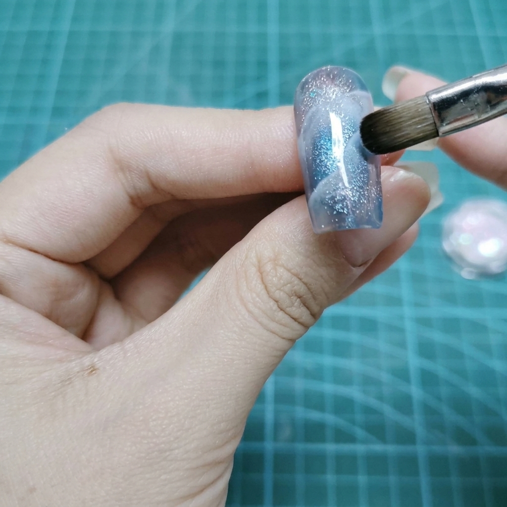 Un pennello da trucco piatto picchietta particelle di glitter olografico sulla superficie bagnata di ununghia in gel.