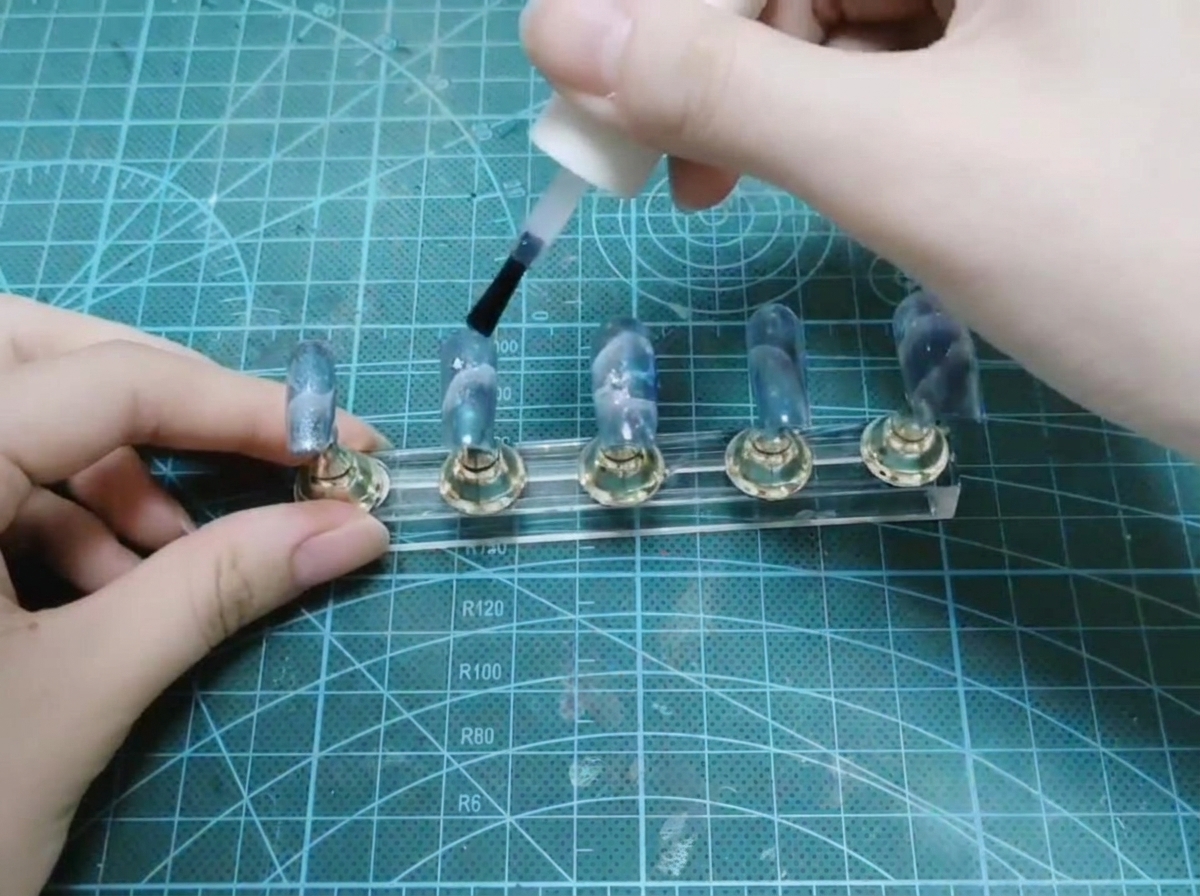 Una mano usa il pennello di una boccetta bianca per applicare top coat lucido su un set di unghie completate su un espositore.