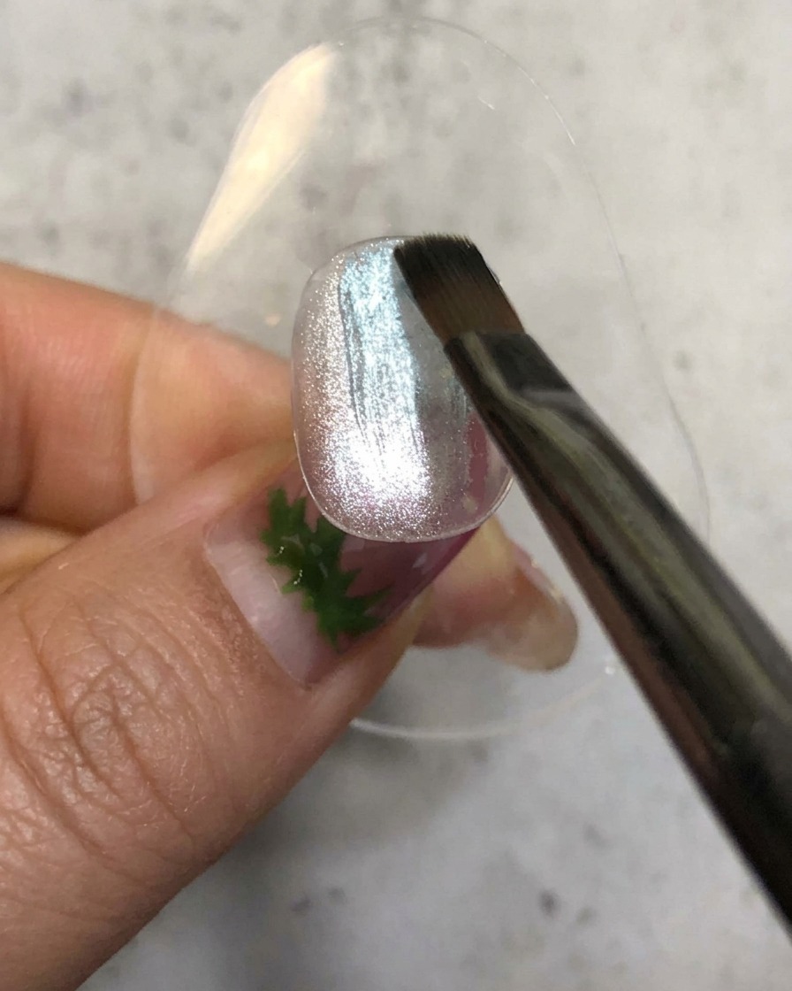 Un pennello da unghie che applica uno smalto gel blu velato su una base magnetica argentata brillante.