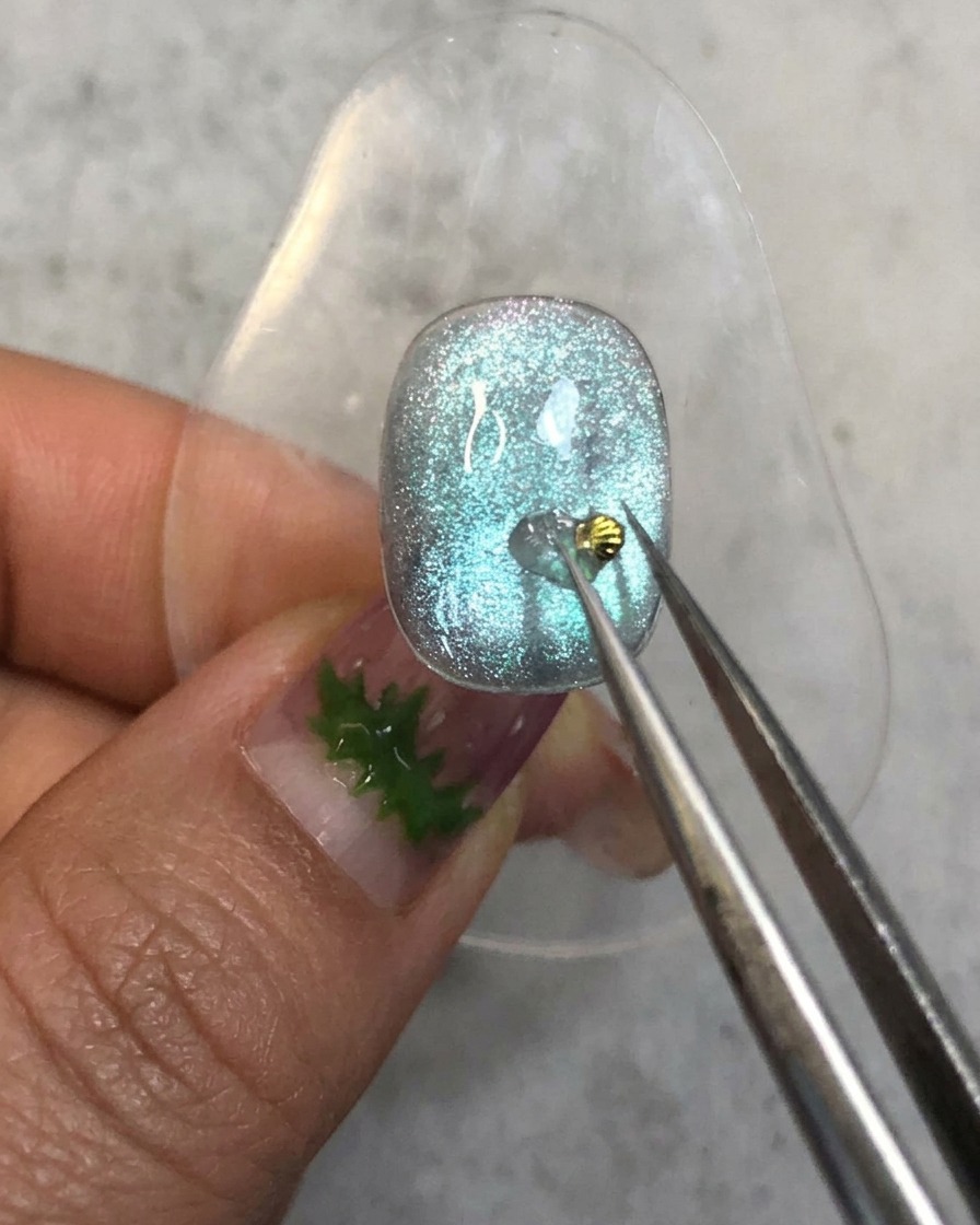 Un paio di pinzette che posizionano un minuscolo charm a forma di conchiglia dorata sulla goccia di gel rinforzante su ununghia blu.