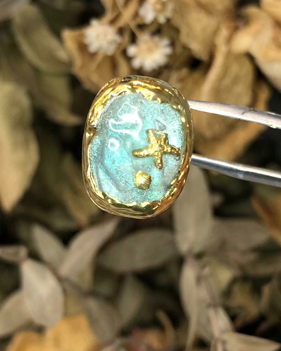 Un paio di pinzette che tengono un pezzo completato di nail art a tema Atlantide con cornice dorata.