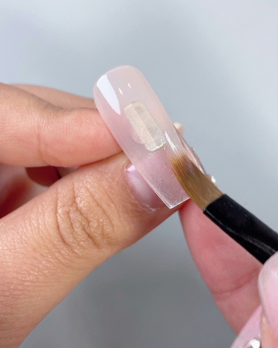 Primo piano di un pennello piatto da trucco che sfuma gel rosa traslucido verso il basso su una tip per unghie finte trasparenti.