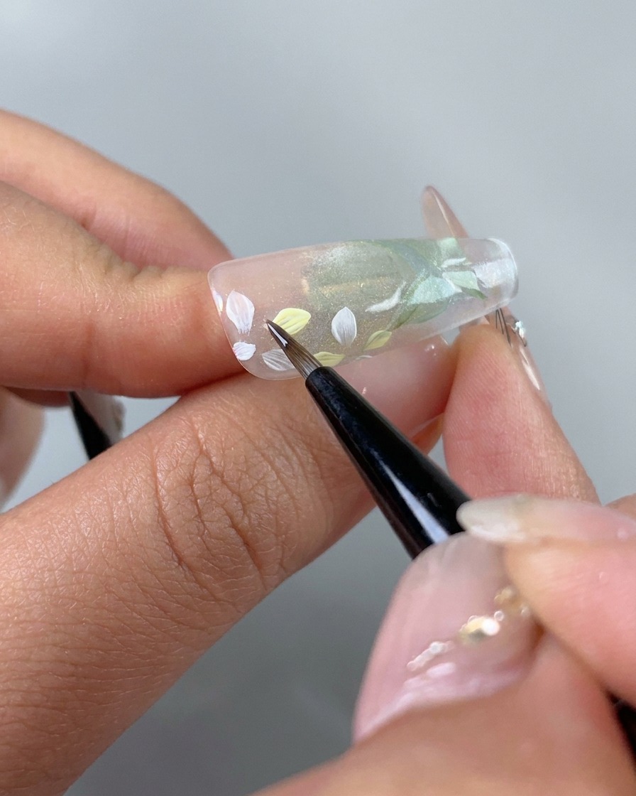 Un pennello fine che aggiunge con attenzione sfumature di gel rosa chiaro e giallo sui singoli petali di fiori dipinti di bianco sullunghia.
