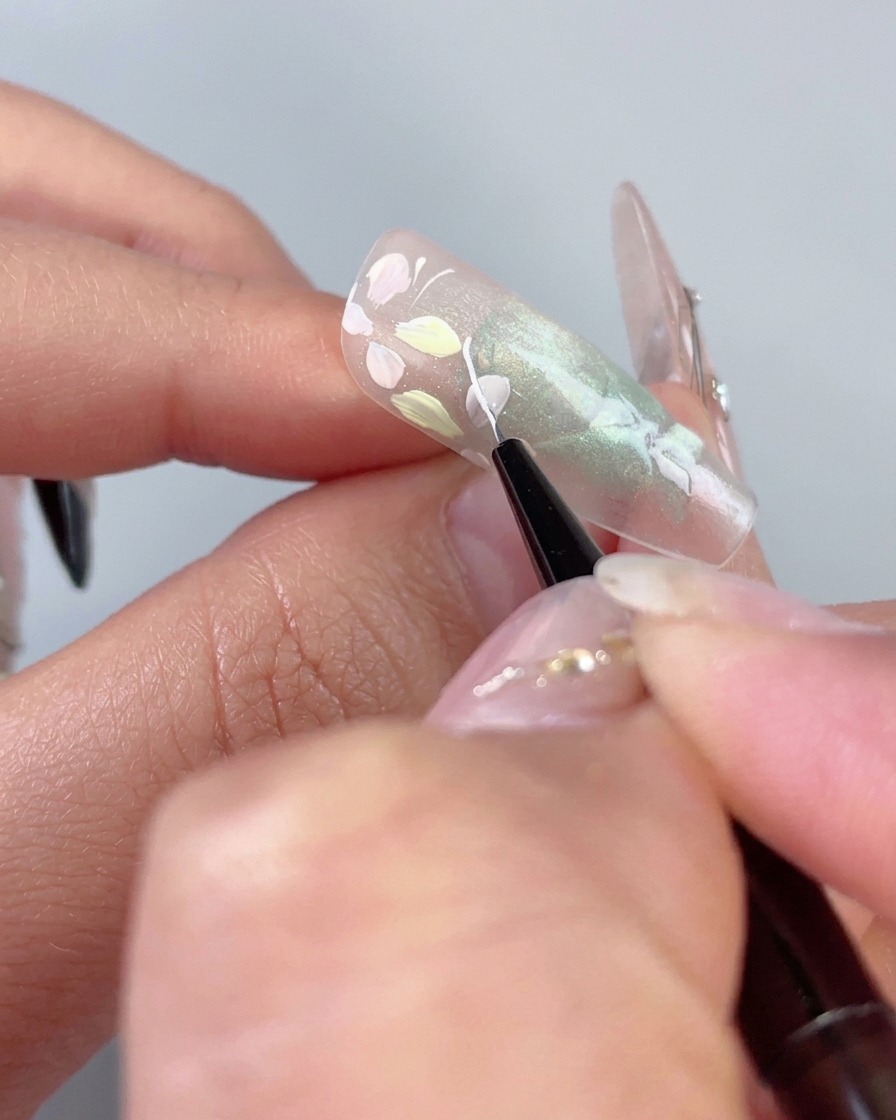 Un pennello liner fine che disegna una delicata linea bianca curva accanto a fiori pastello su ununghia verde trasparente cat-eye.