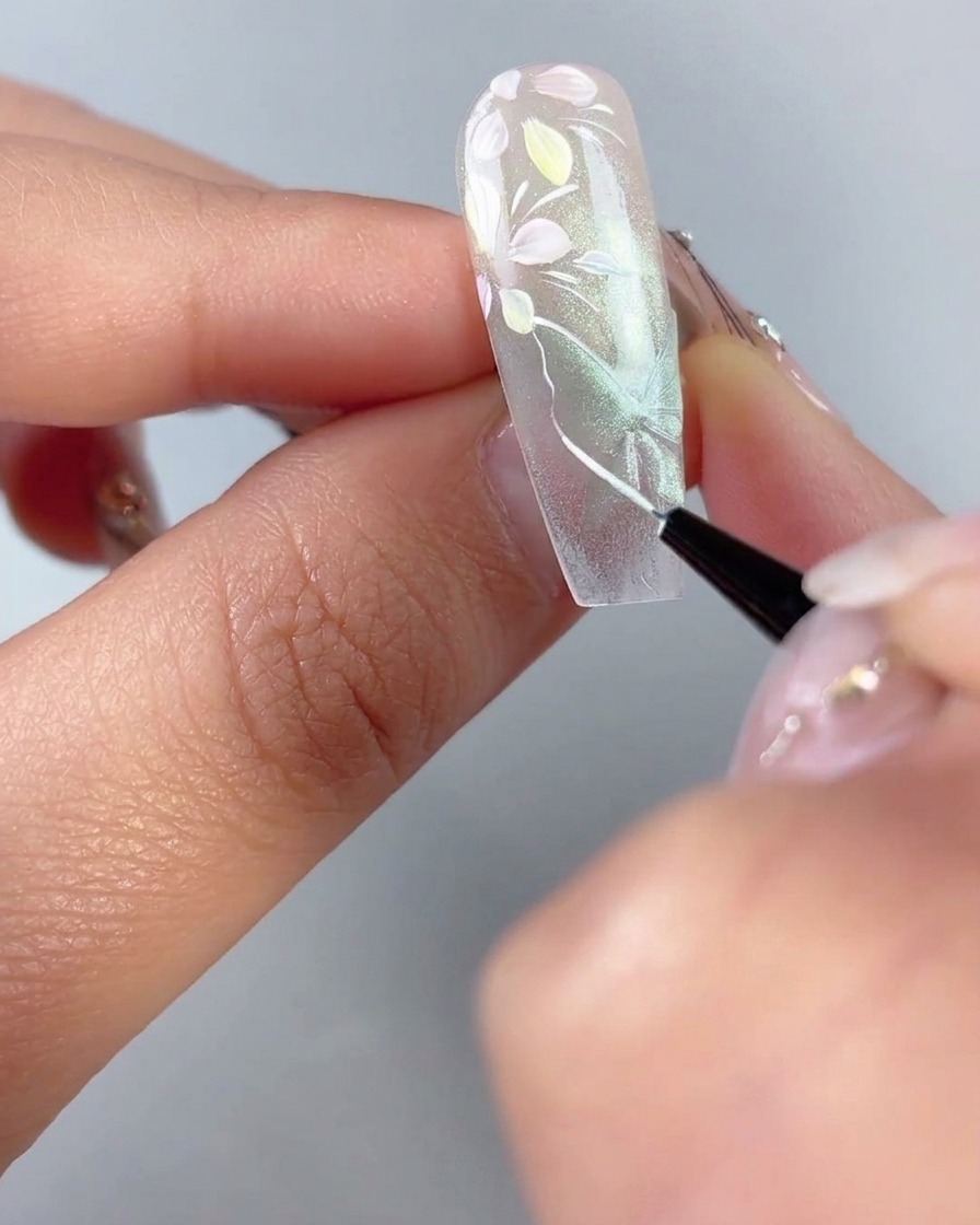 Un pennello liner fine che traccia sottili contorni bianchi e venature delle ali su una forma di farfalla verde trasparente cat-eye su ununghia.