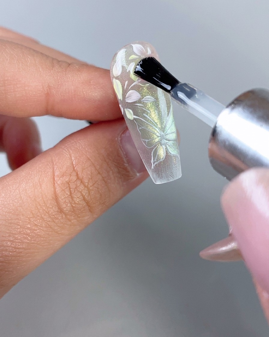 Un pennello per smalto che applica un top coat trasparente lucido su ununghia trasparente con decorazioni floreali e di farfalle completamente rifinite.