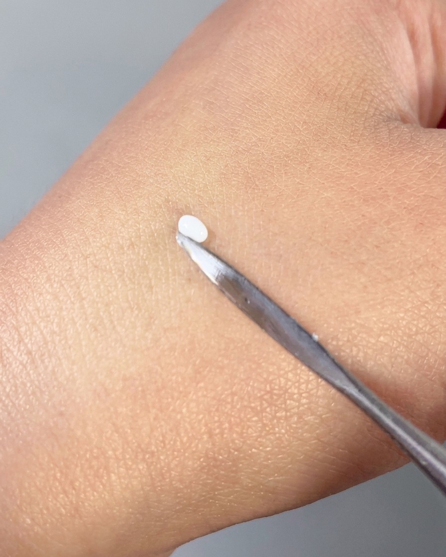 Una spatola di metallo che preme una piccola goccia di gel 3D bianco in una forma di petalo liscia sul dorso di una mano.