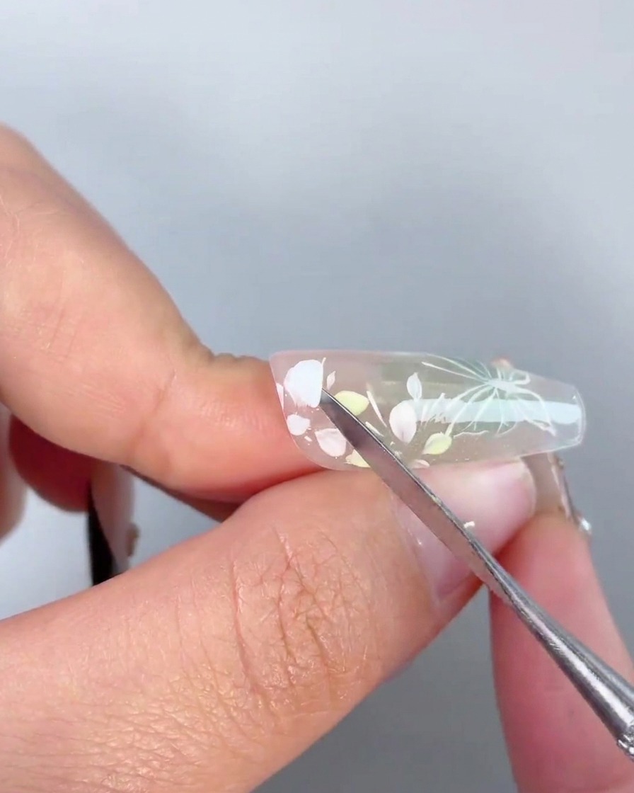 Uno strumento di metallo che applica una goccia di gel adesivo trasparente al centro di un design floreale dipinto su ununghia.
