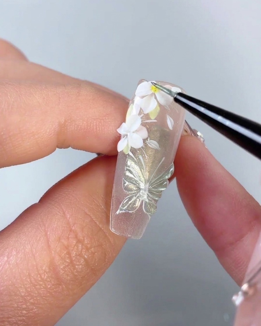 Un pennello fine che aggiunge un minuscolo punto giallo al centro di un fiore bianco 3D su ununghia.