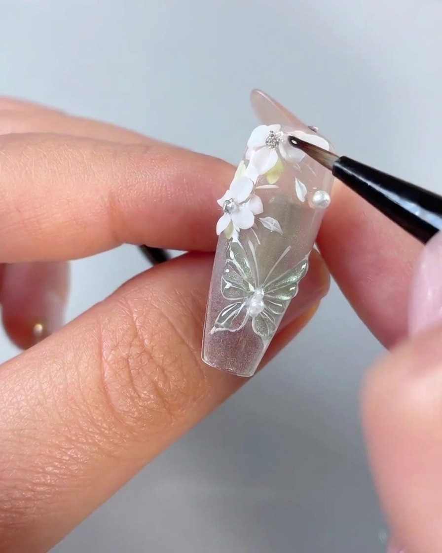 Un pennello di dettaglio fine per unghie che applica gel trasparente attorno ai bordi dei petali di fiori 3D bianchi e strass su una tip trasparente.