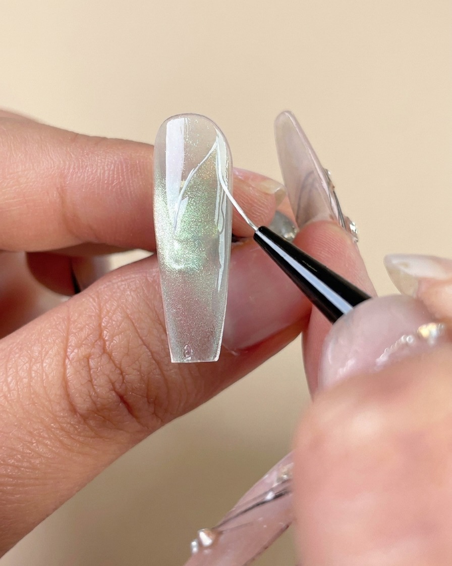 Un pennello liner fine che disegna linee di venature bianche su ununghia in gel cat-eye verde.