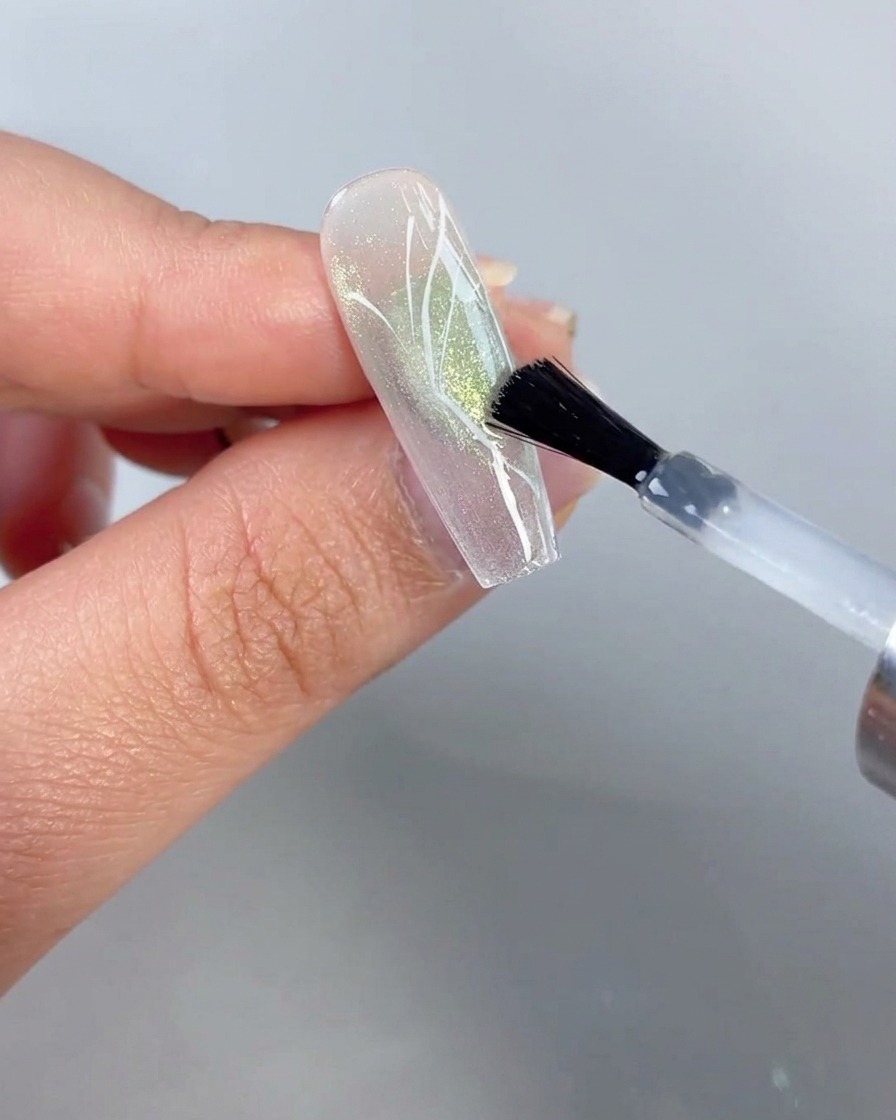 Un pennello che applica un top coat trasparente su ununghia con un design a mezza farfalla dipinto su uno sfondo cat-eye verde trasparente.