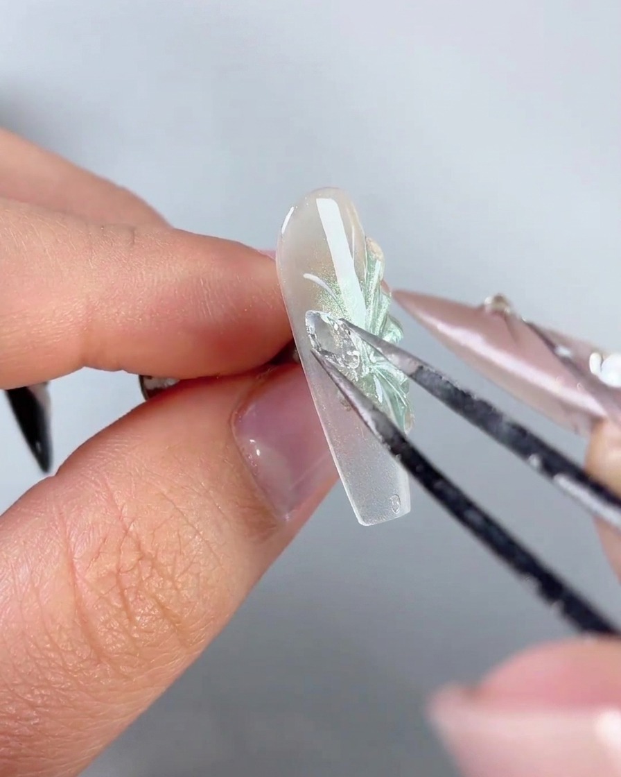 Un paio di pinzette di metallo che posizionano uno scintillante ciondolo ad ala di farfalla in strass sul lato di ununghia decorata.