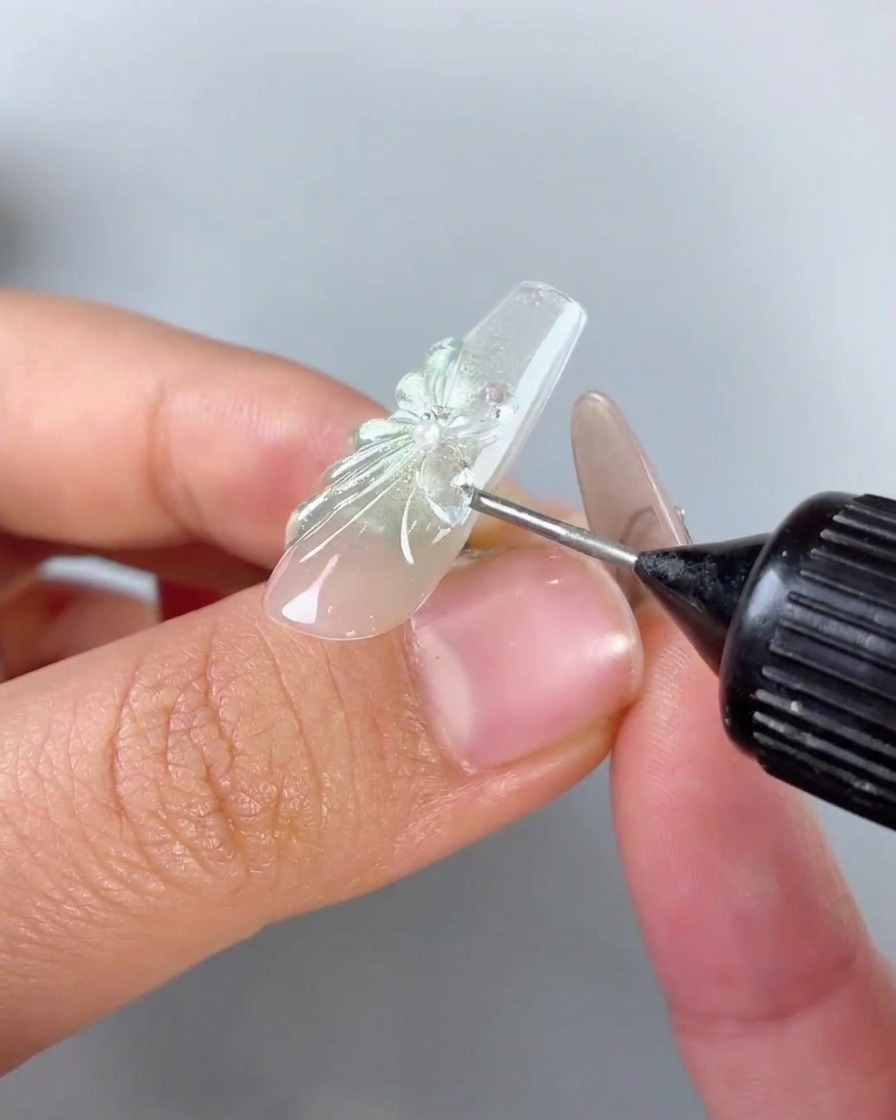 Un flacone con beccuccio di precisione che applica gel riempitivo trasparente attorno alla base e ai bordi di un ciondolo a farfalla in strass su ununghia.