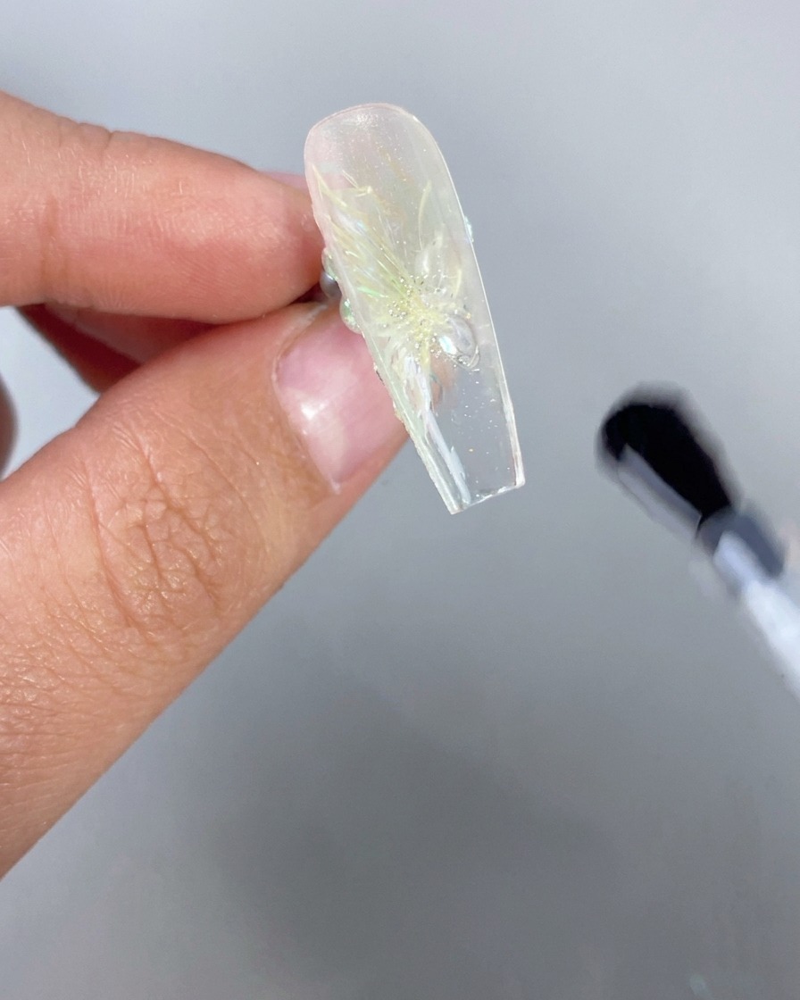 Primo piano di una mano che tiene capovolta ununghia trasparente effetto ghiaccio con un design floreale 3D, mentre un pennello applica il top coat trasparente sulla parte inferiore del bordo libero.