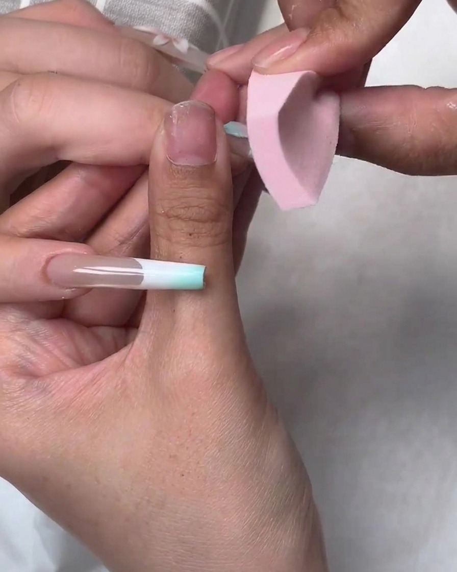 Una piccola spugnetta rosa tampona lo smalto gel blu sui bordi di unapplicazione ungueale durante un tutorial di nail art.