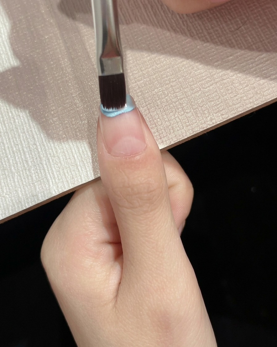 Un pennello piatto che applica lo smalto gel azzurro sulla metà superiore della piastra ungueale.