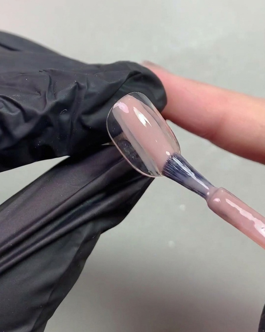 Primo piano di una mano con guanto nero che usa un pennello per applicare un gel nude trasparente dai toni aranciati su una tip.