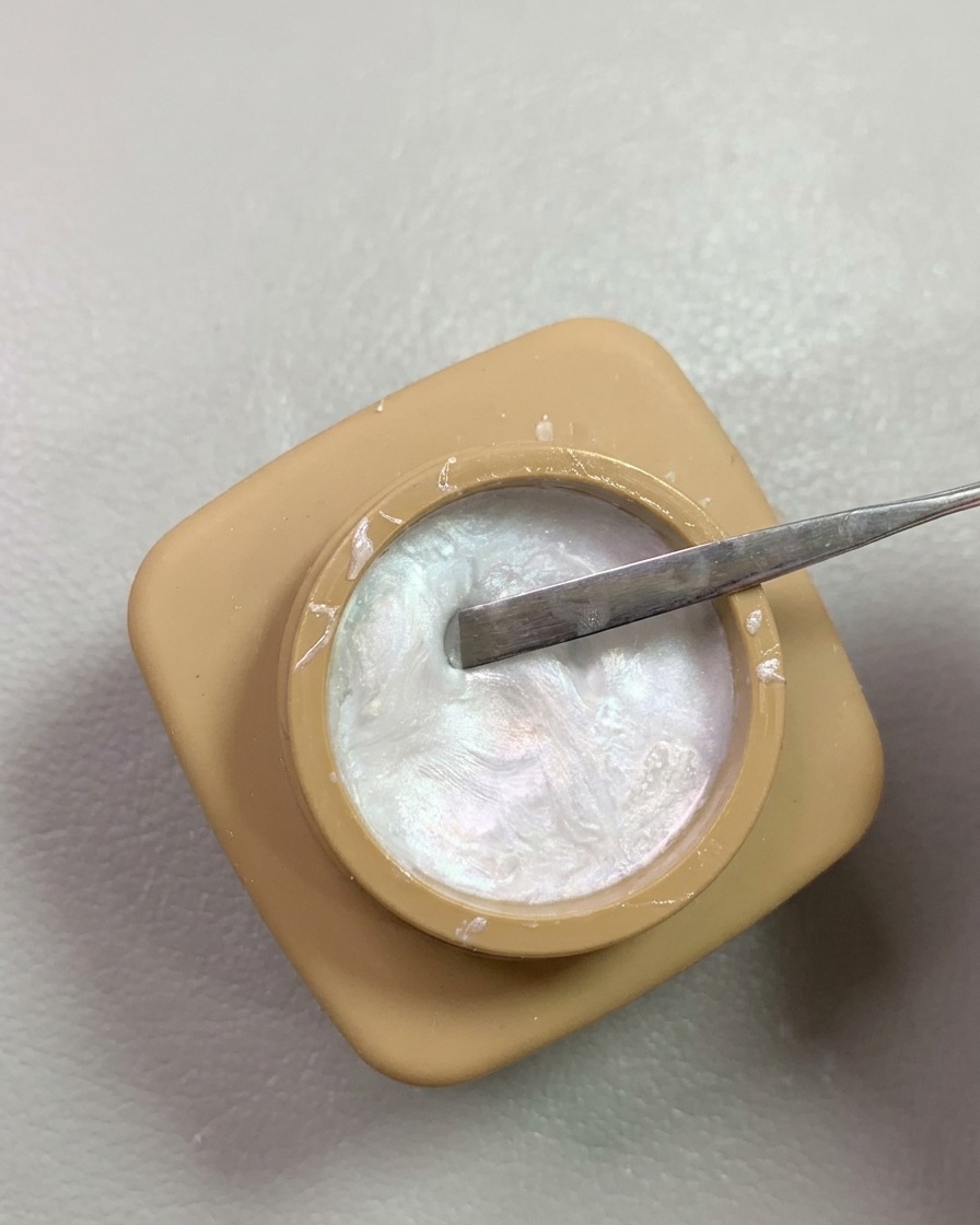 Una spatola di metallo che preleva gel da scultura perla madreperla brillante da un piccolo barattolo color sabbia.