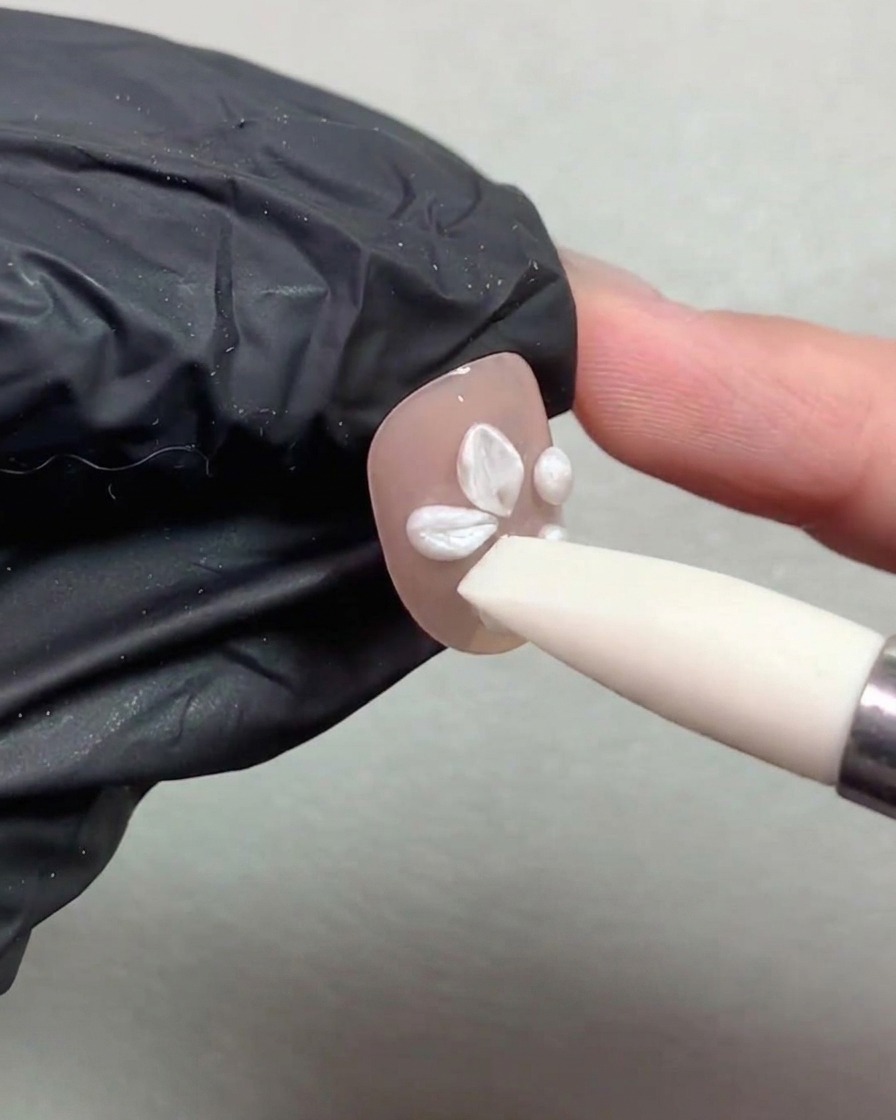 Uno strumento penna in silicone che preme e modella sfere di gel perla bianca in petali di fiore su ununghia dipinta color nude.