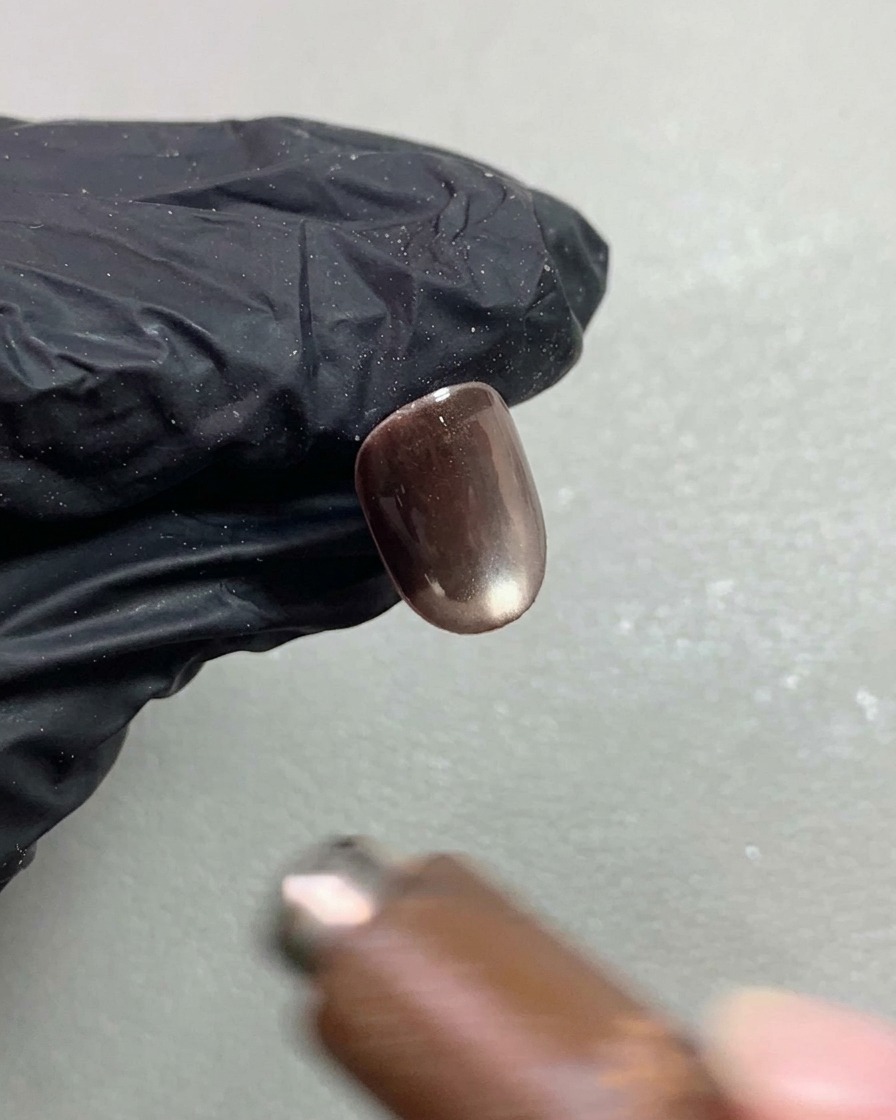 Un magnete per unghie che si libra sopra una corta unghia in gel cat-eye marrone scuro, modellando il pigmento riflettente in un effetto sferico luminoso.