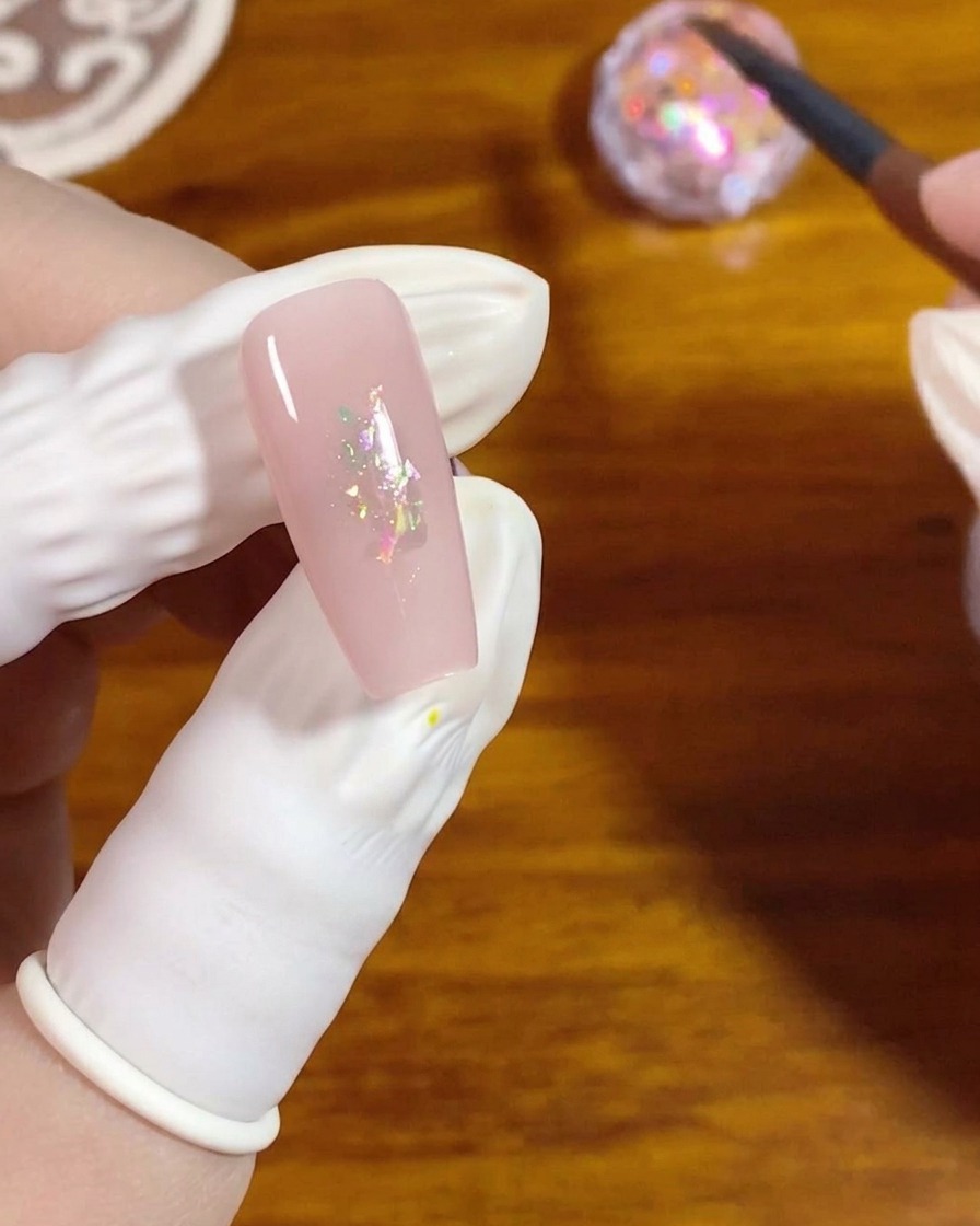 Un pennello piatto per gel preme piccole scaglie opalescenti cangianti al centro di ununghia smaltata di rosa.