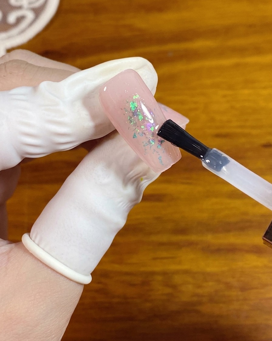 Un pennello per gel applica un top coat opaco trasparente su ununghia rosa con scaglie opalescenti al centro.