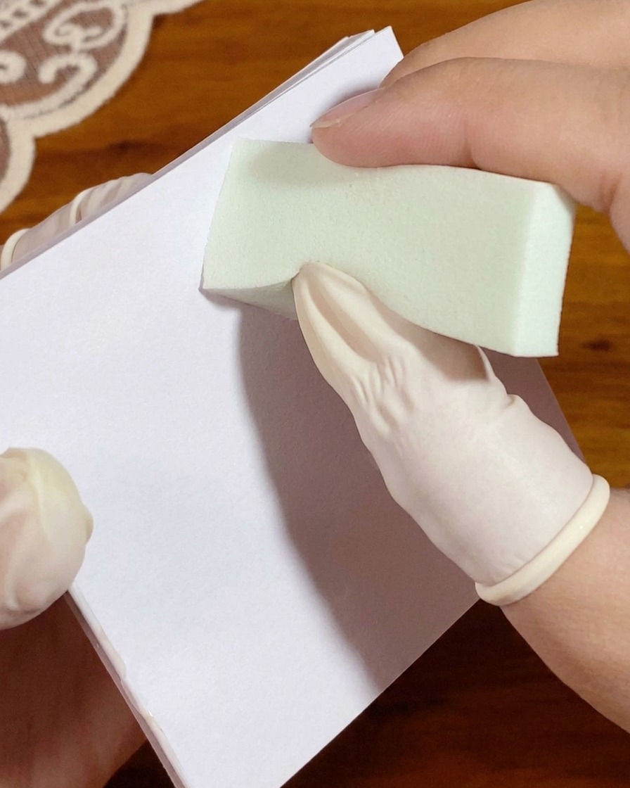 Una mano con i guanti preme una spugnetta carica di gel bianco su una carta da tavolozza prima dellapplicazione sullunghia.