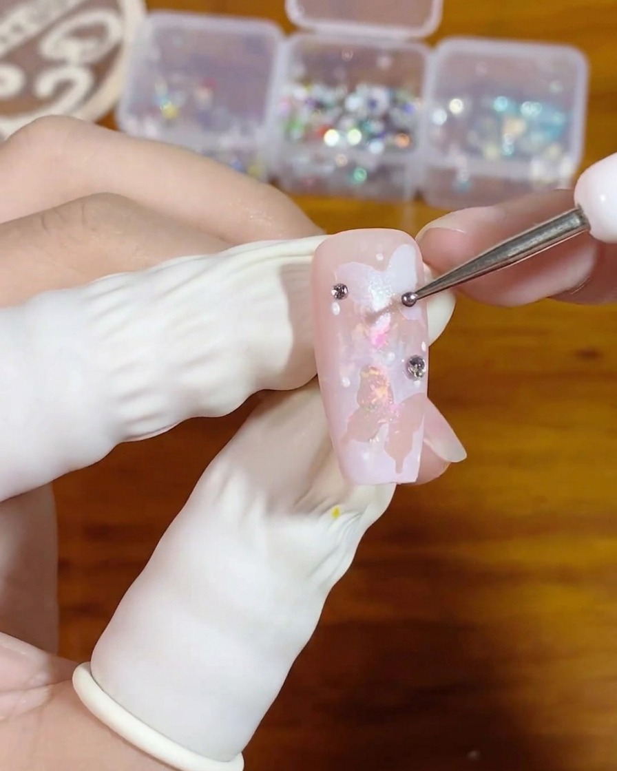 Primo piano di una mano che usa un dotting tool sottile per posizionare una piccolissima goccia di gel trasparente su ununghia rosa decorata con farfalle bianche.