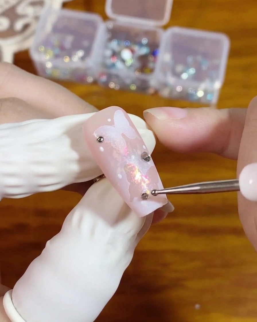 Un dotting tool posiziona un piccolo strass argentato a base piatta su una goccia di gel trasparente su ununghia rosa con un design a farfalla.