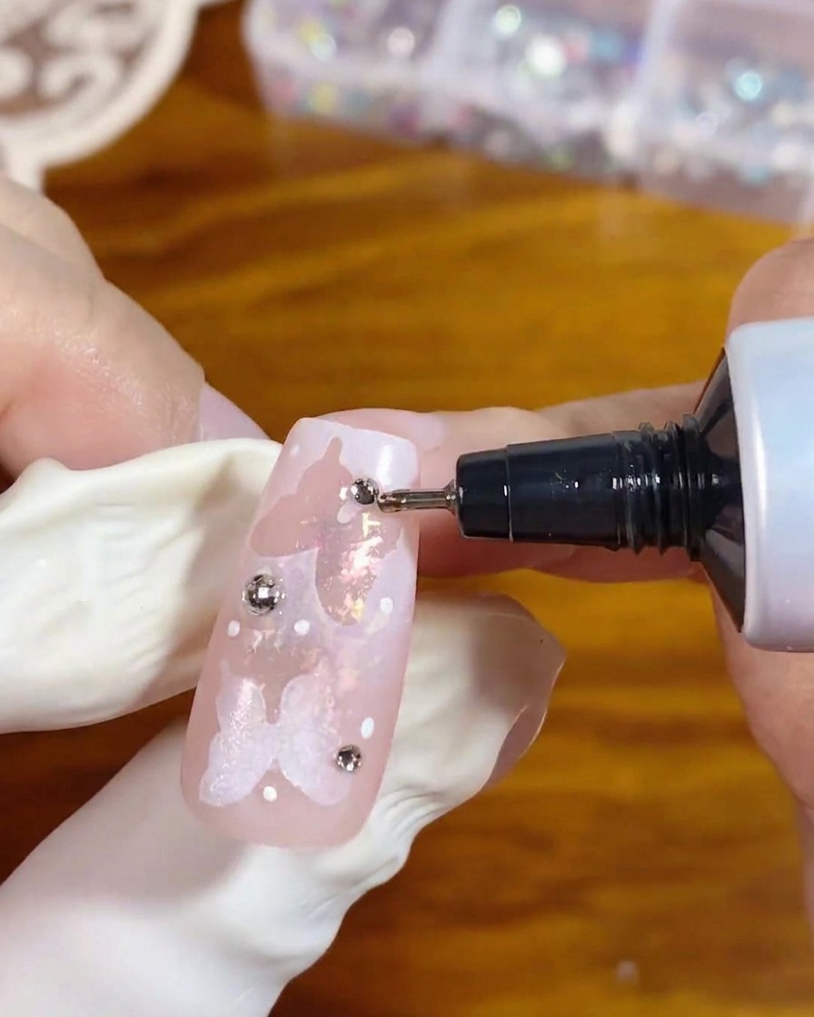 Un tubetto applicatore di precisione distribuisce del gel trasparente attorno alla base di uno strass argentato su ununghia rosa decorata per fissarlo.