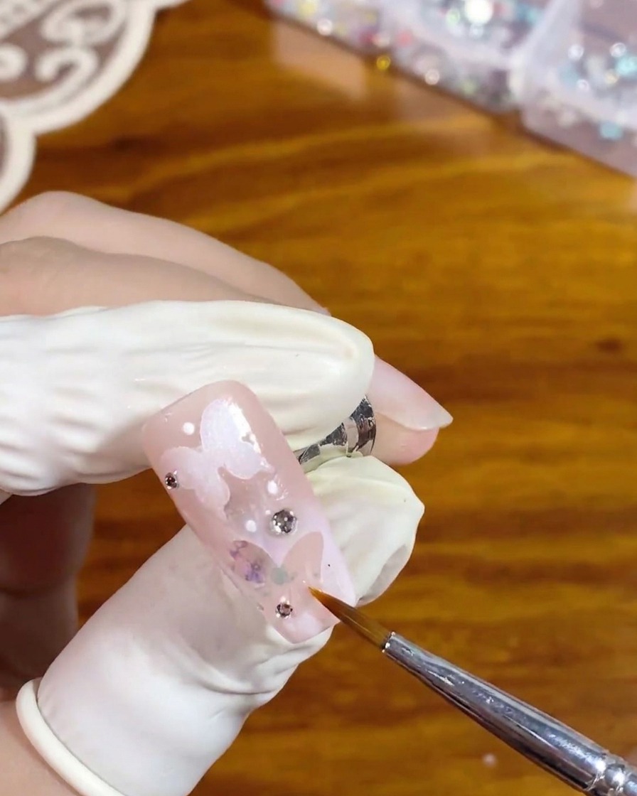 Un pennello liner sottile applica con cura un top coat lucido trasparente attorno a uno strass argentato su ununghia rosa con un design a farfalla bianca in negativo.