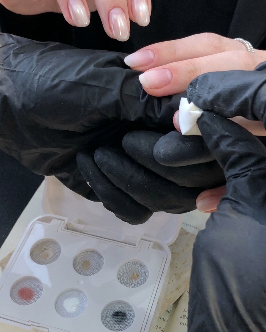 Una mano guantata che utilizza uno strumento a spugna bianco per applicare una sfumatura bianco avorio su ununghia vicino alla linea delle cuticole.